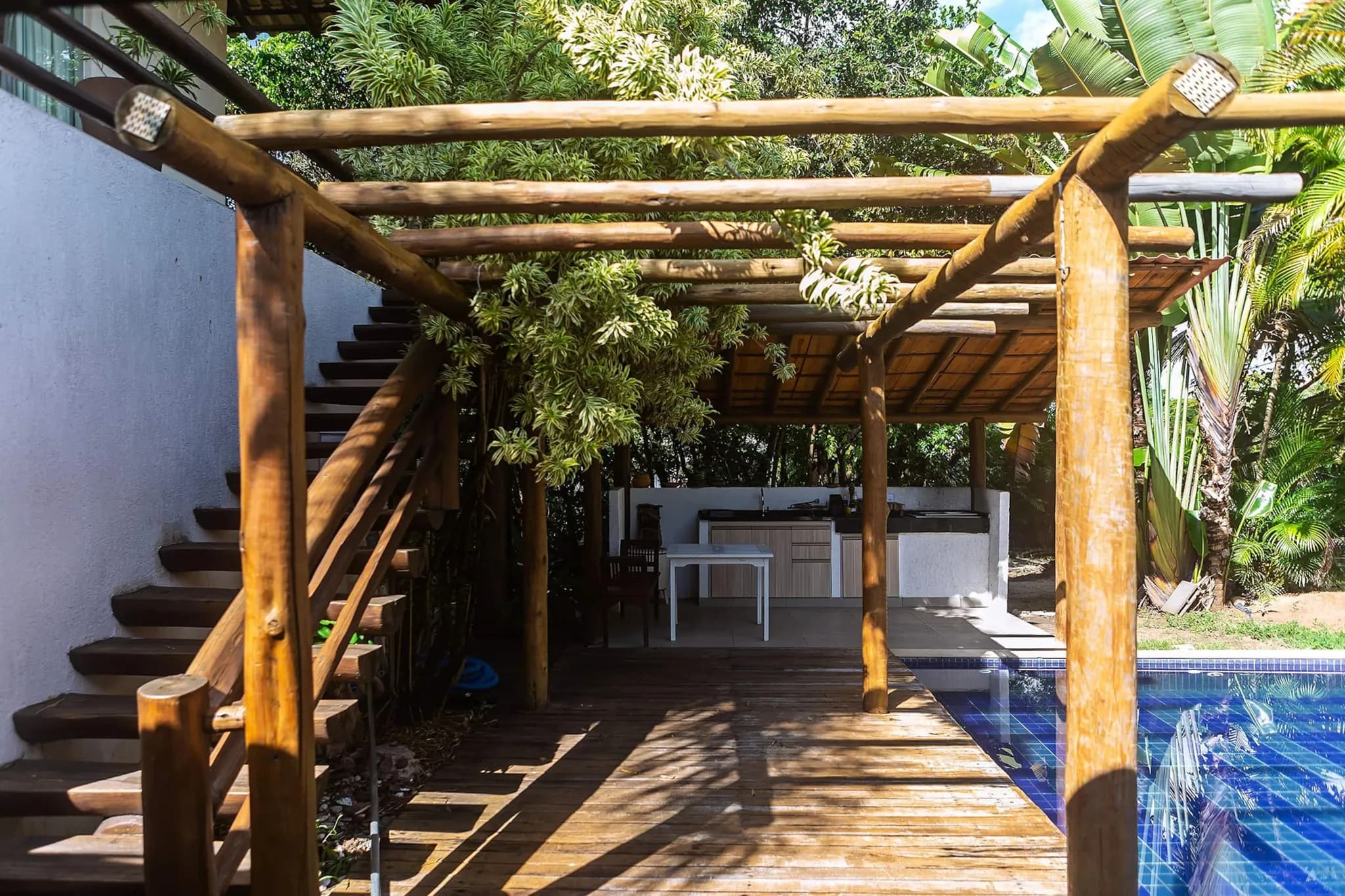 Pergolado de madeira com escada, a área da churrasqueira ao fundo e a piscina à direita
