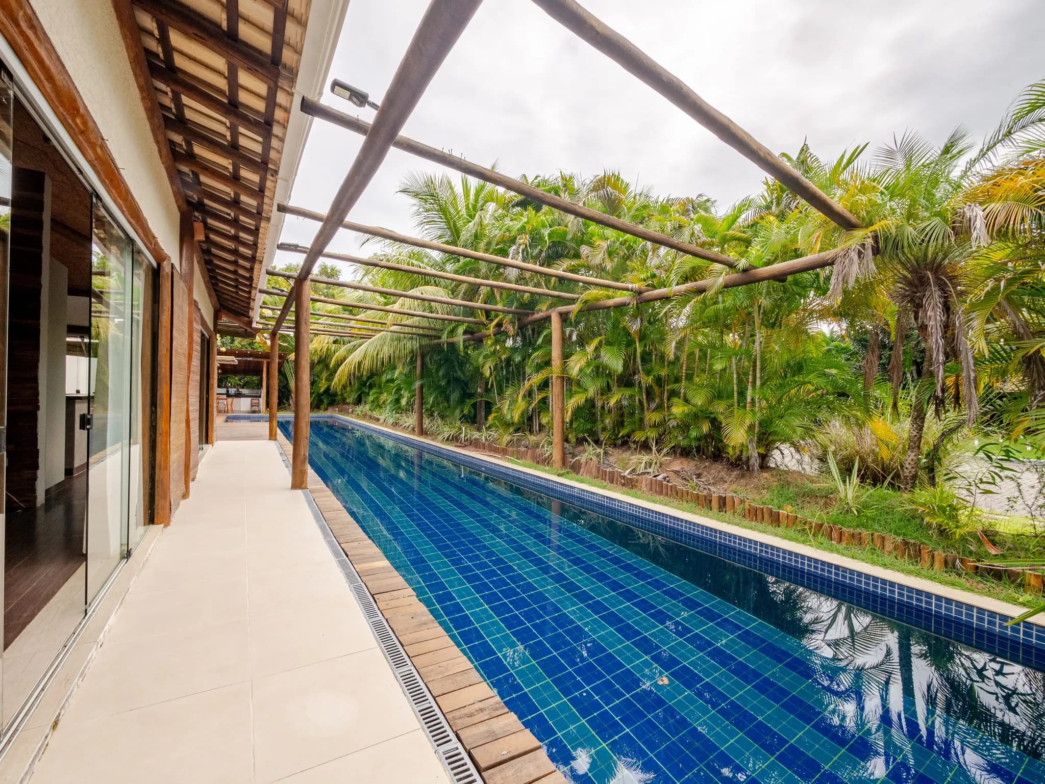 Raia da piscina ao lado da varanda da casa, com jardim de palmeiras do outro lado