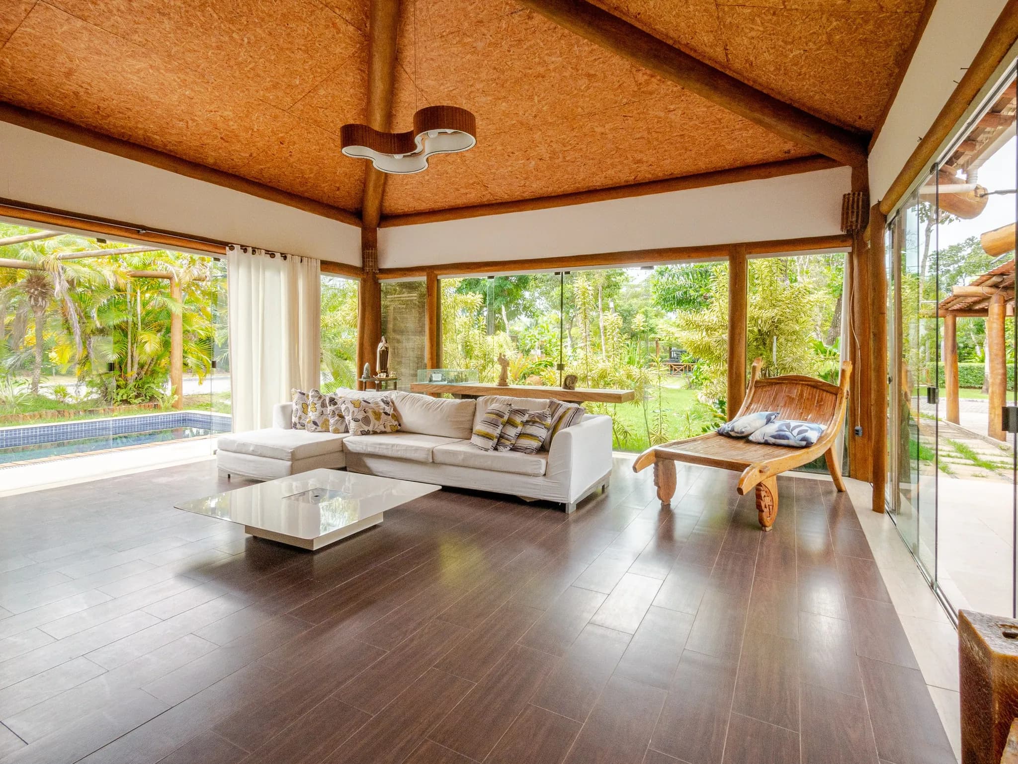 Sala de estar com sofá branco, piso escuro e vidros do chão ao teto abertos para o jardim e a piscina