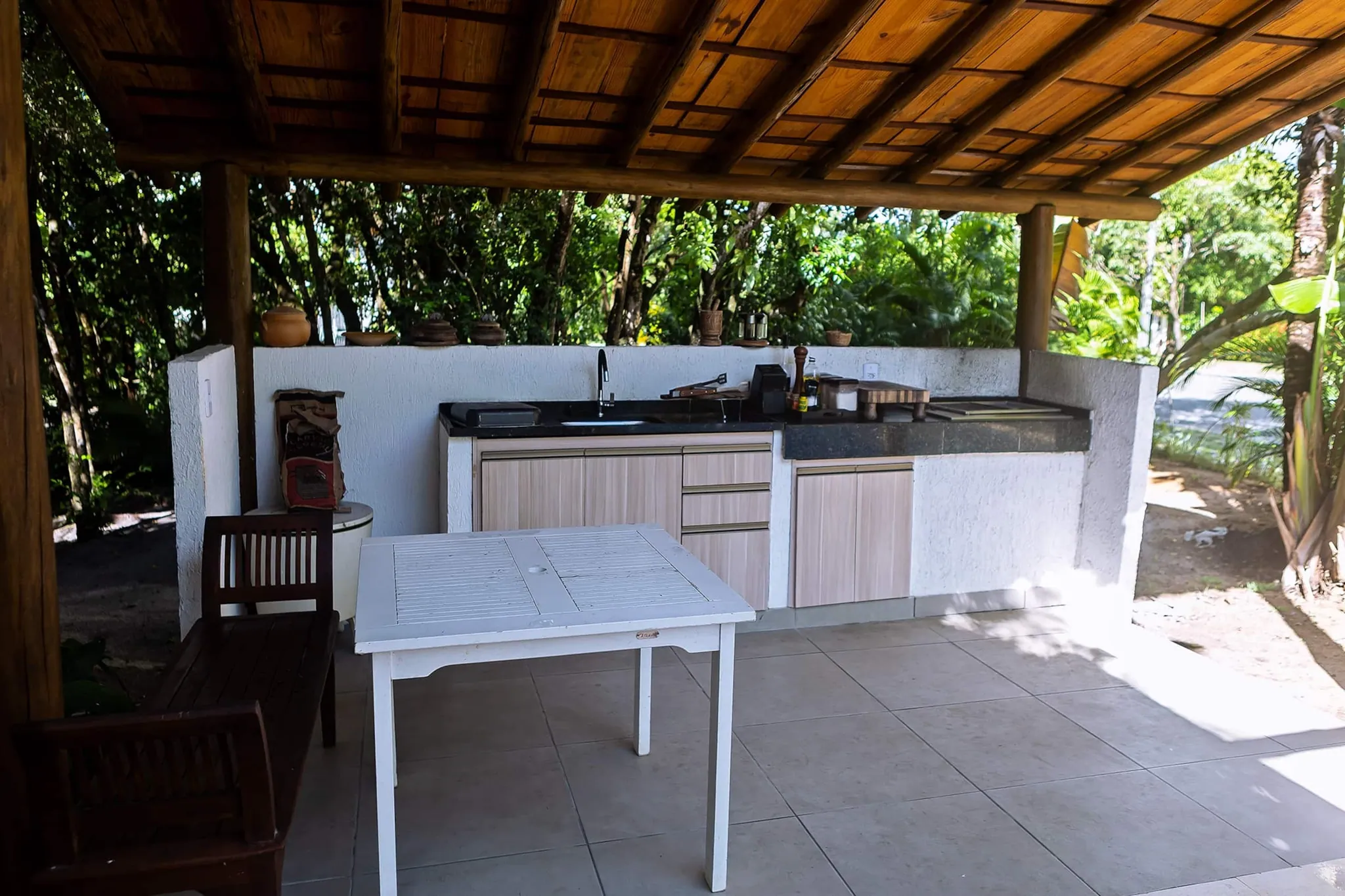 Churrasqueira externa com bancada, pia e mesa branca sob cobertura de madeira