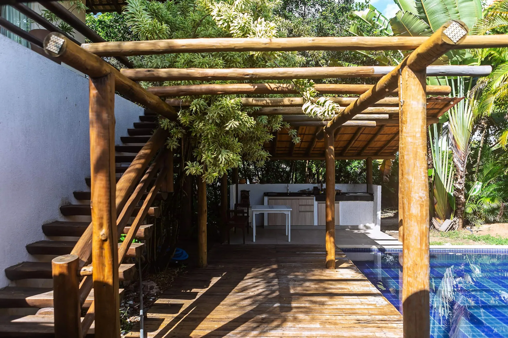 Pergolado de madeira com escada, a área da churrasqueira ao fundo e a piscina à direita