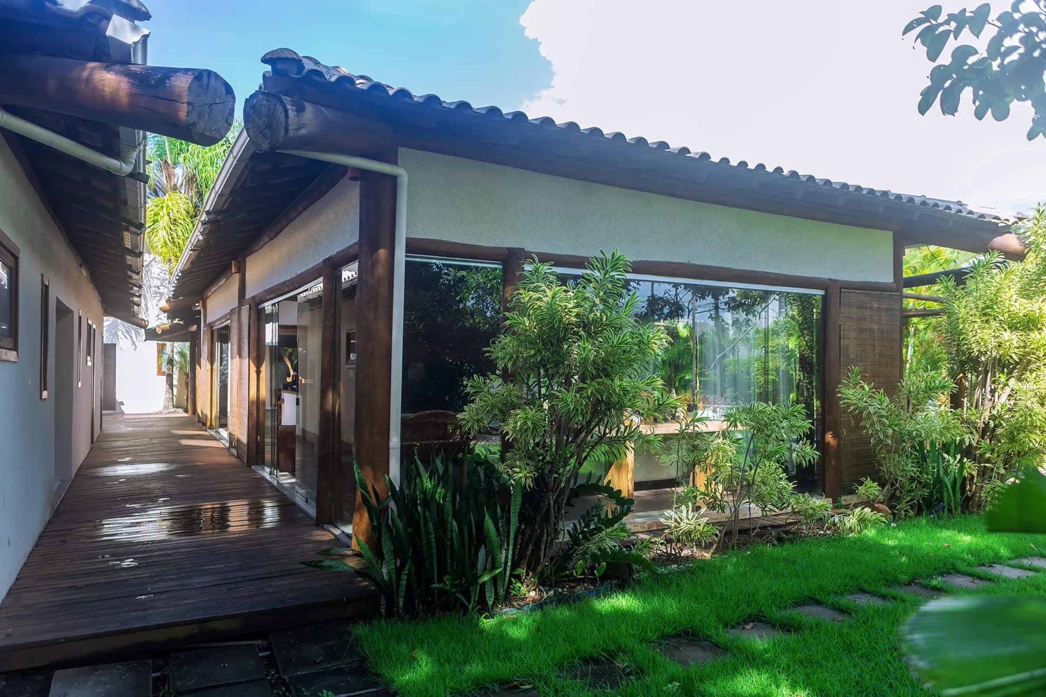 Lateral da casa com passarela de madeira, janelões e gramado com plantas