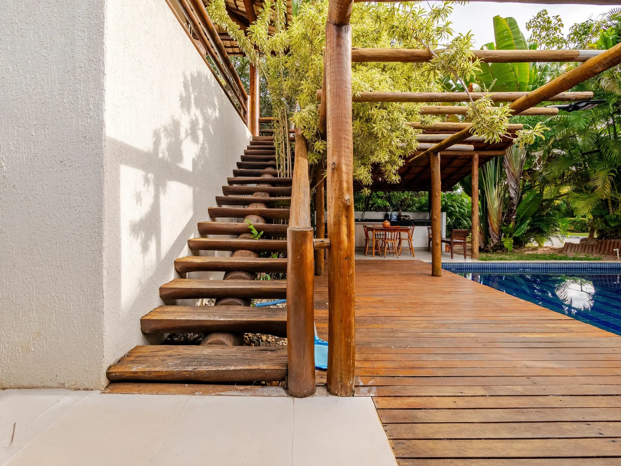 Escada de madeira que desce ao deck, com pergolado coberto de folhas e piscina ao fundo