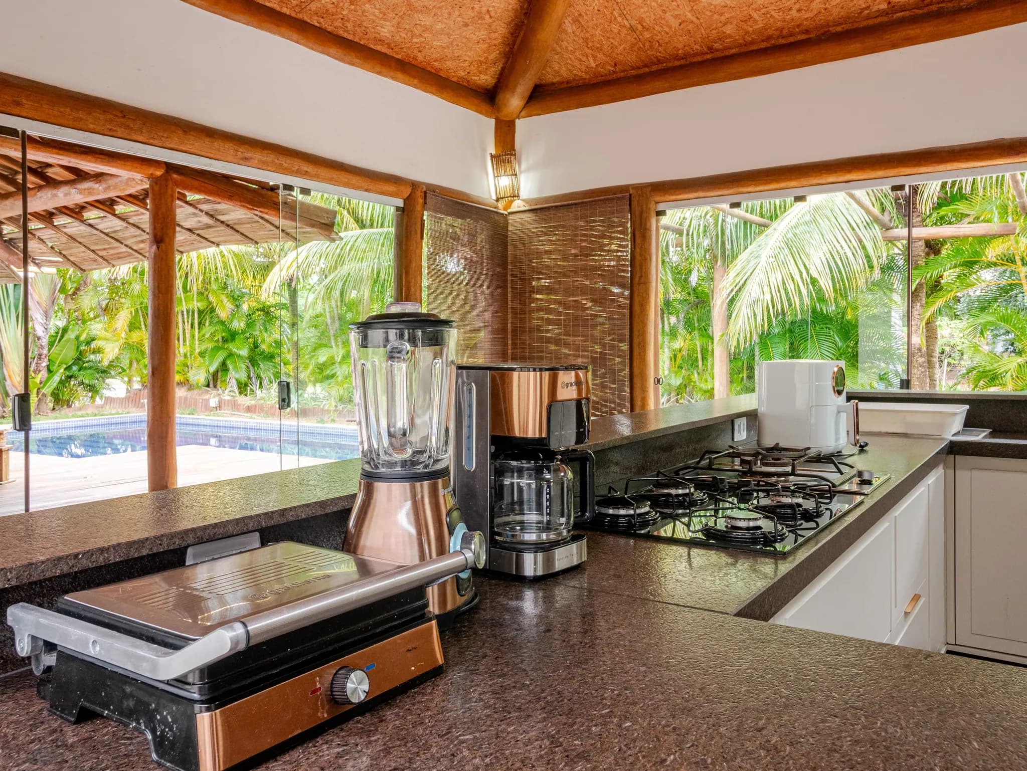 Bancada da cozinha com cooktop, liquidificador e cafeteira, com a piscina vista pela janela