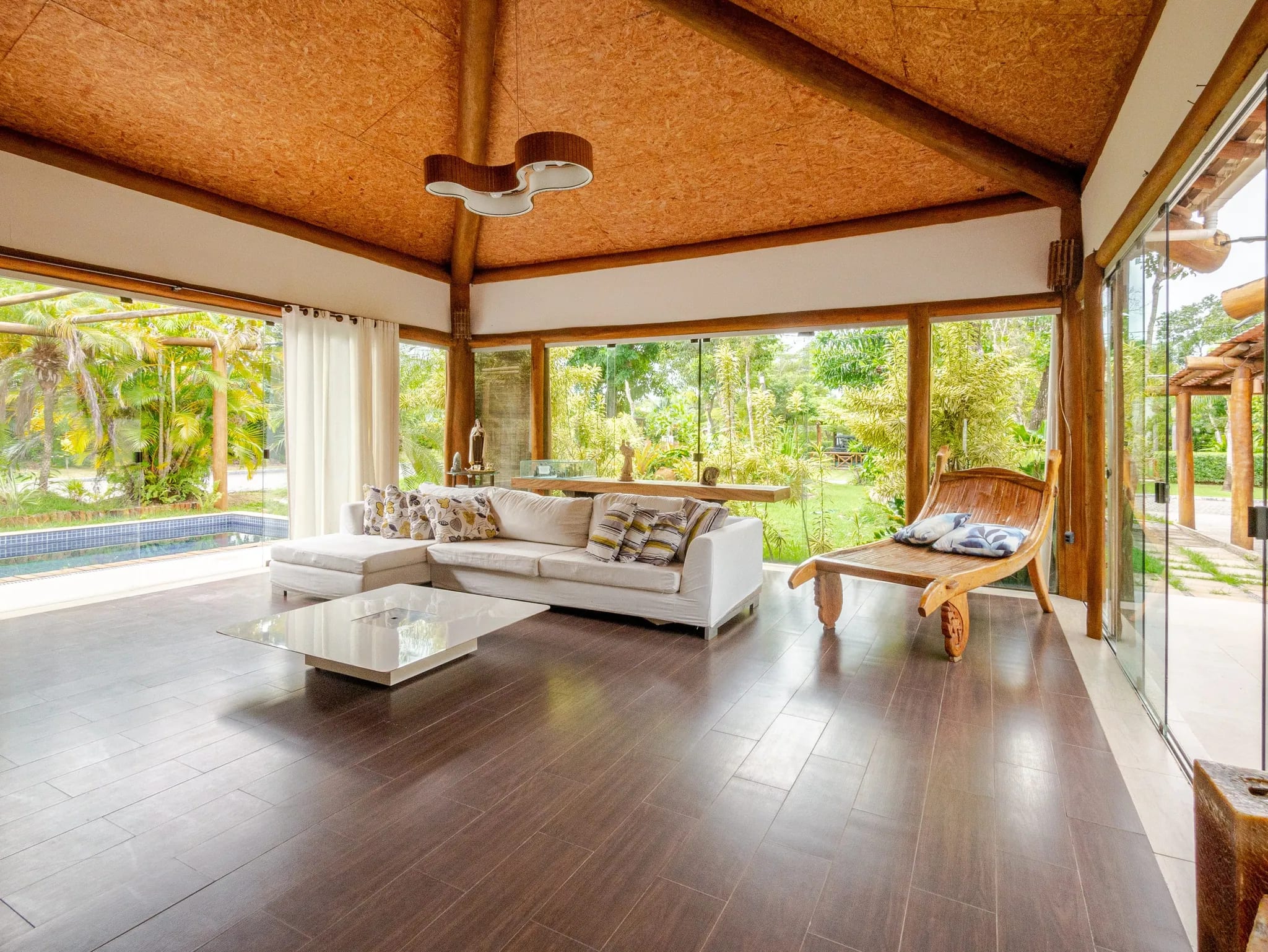 Sala de estar com sofá branco, piso escuro e vidros do chão ao teto abertos para o jardim e a piscina
