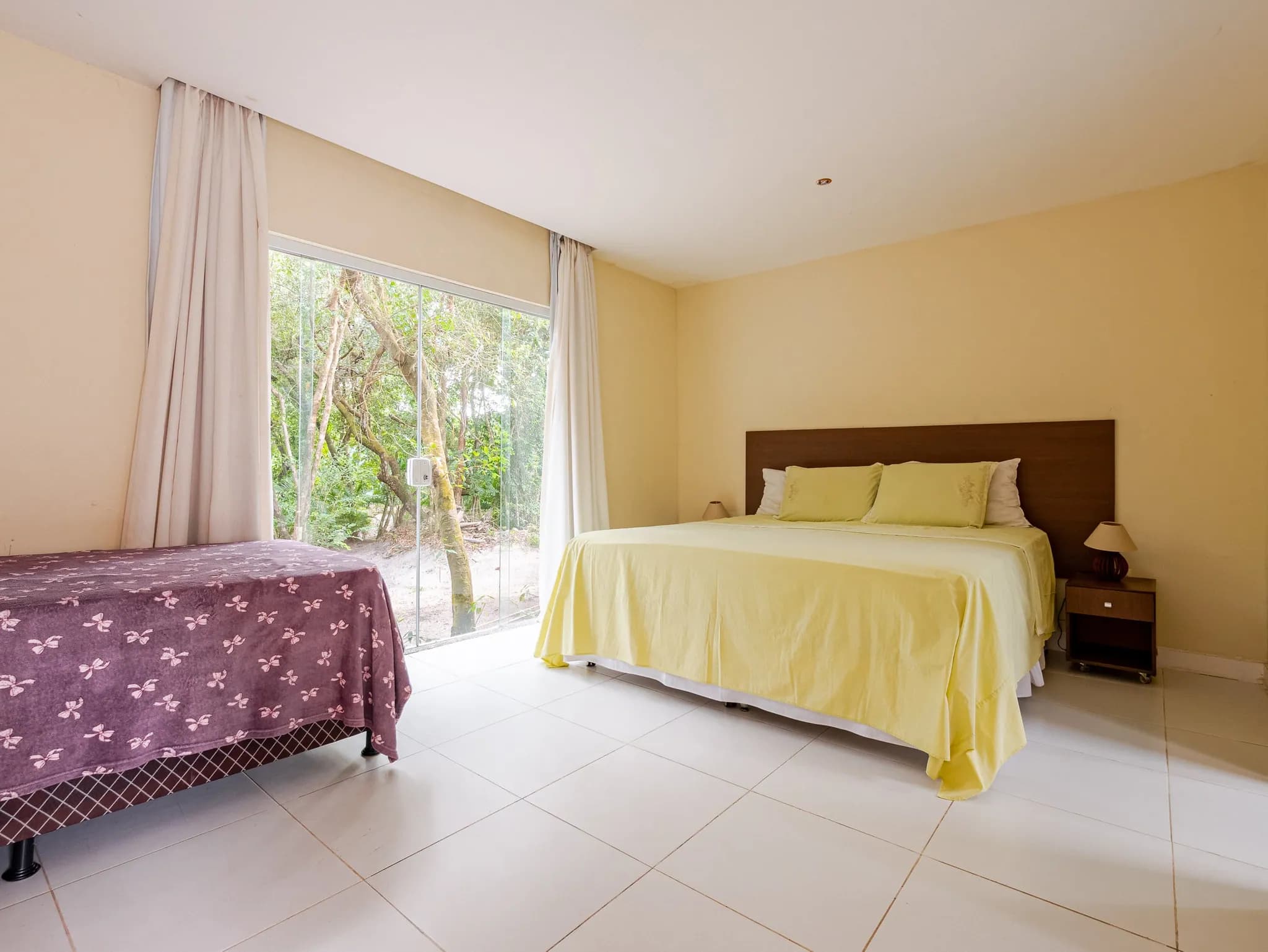 Suíte com cama de casal e cama de solteiro diante de janela de vidro para a mata